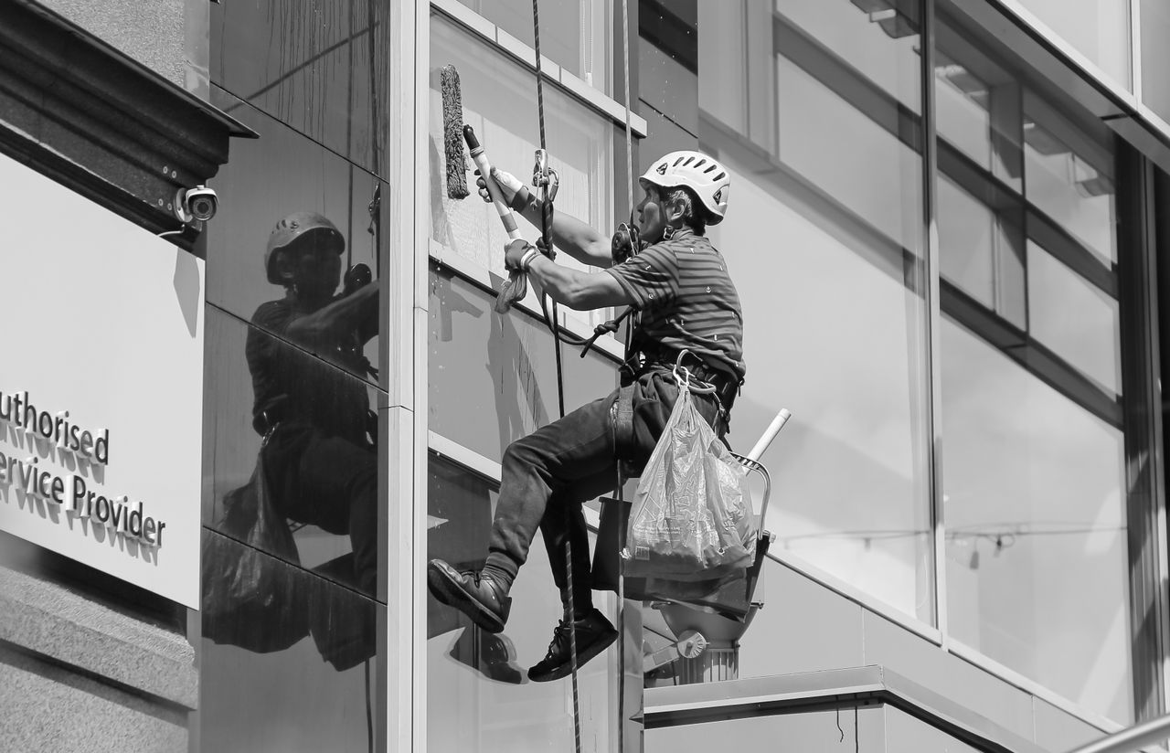 Noir et blanc, vu d’en bas, d’un homme cordiste en casque de protection et équipé de matériel professionnel nettoyant la façade vitrée d’une tour moderne